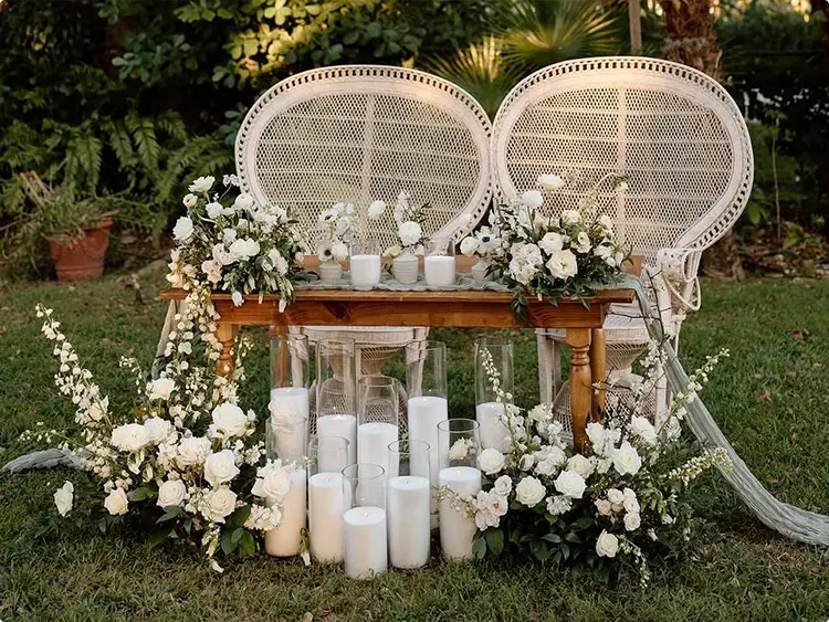 White Roses and Greenery, Two Large White Rattan Peacock Chairs, a Wood Sweetheart Table, and a Tall Glass Pillar Candle