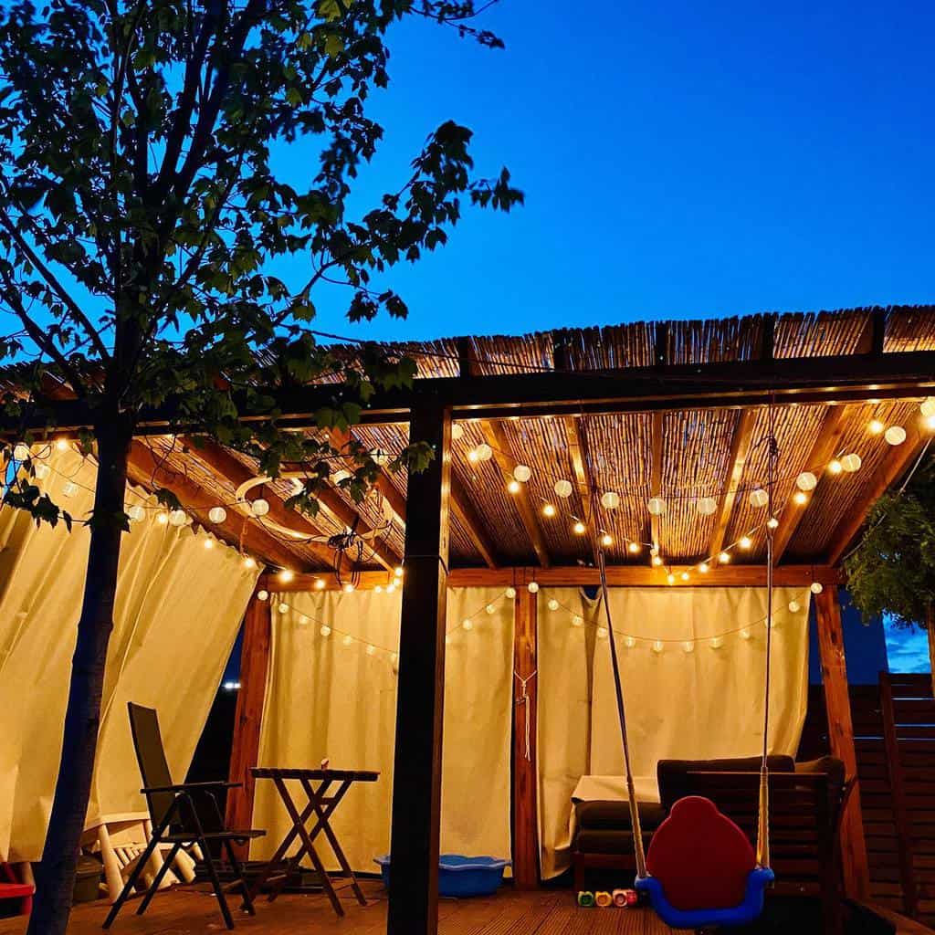 backyard-patio-lighting-ideas-makeitmemorable-ro_ wood deck patio pergola with string lights and curtains