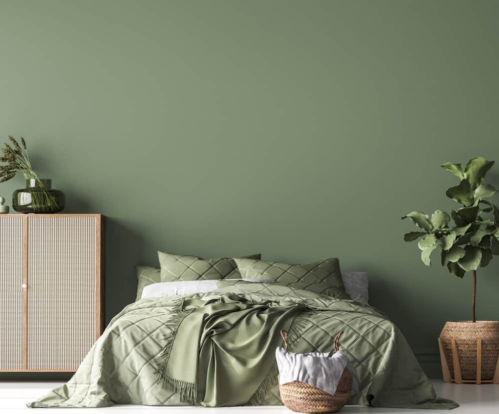 classic-green-bedroom-ideas-3 A green bedroom with a neatly made bed, plants, and a wicker basket on a wooden floor