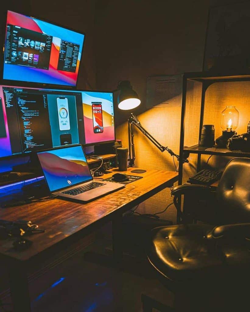 Cozy workspace with a wooden desk, multiple screens, a laptop, and a warm lamp light creating an inviting atmosphere