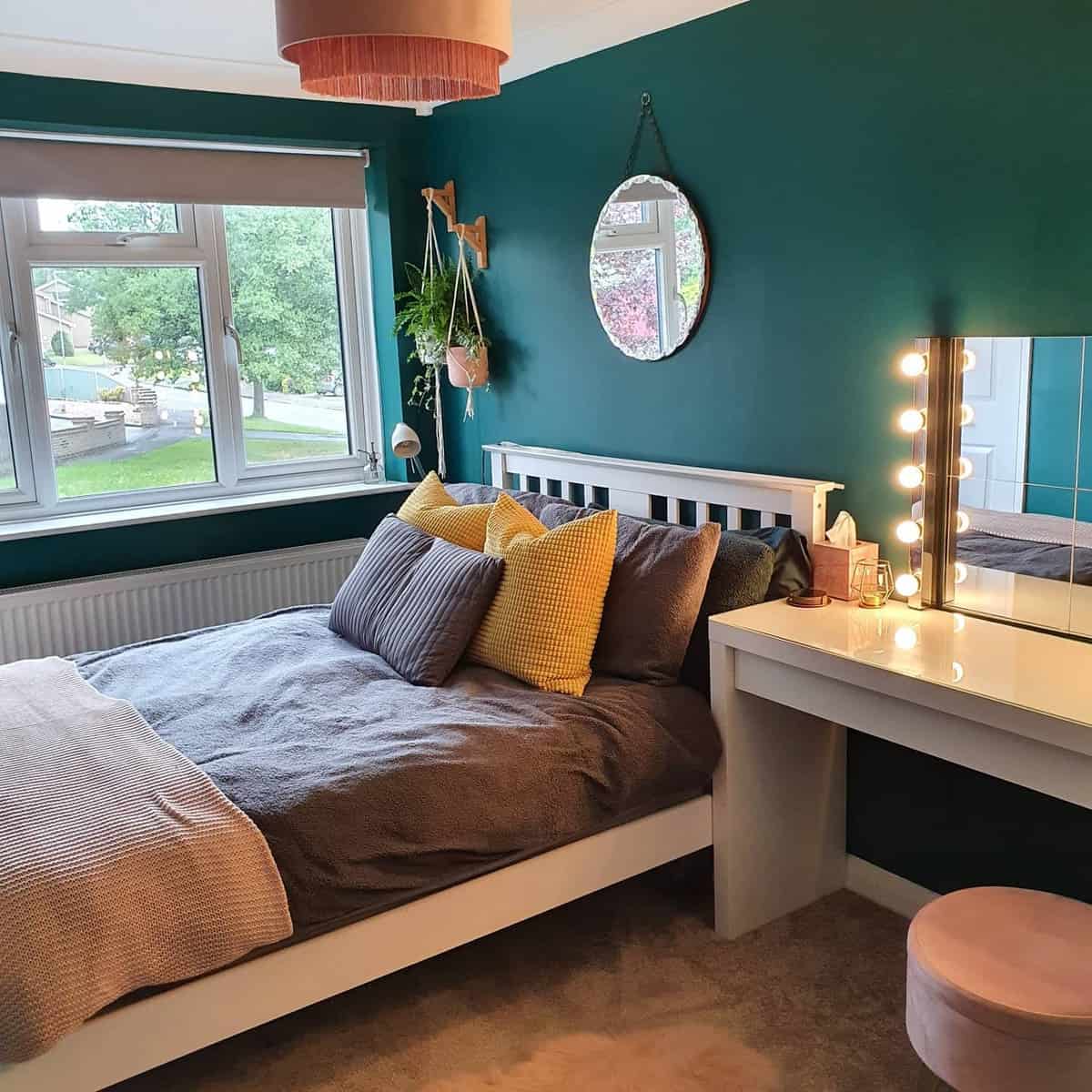 girl-green-bedroom-ideas-my_1970s_home_makeover Bedroom with teal walls, a bed with gray and yellow pillows, a round mirror, and a makeup vanity by the window