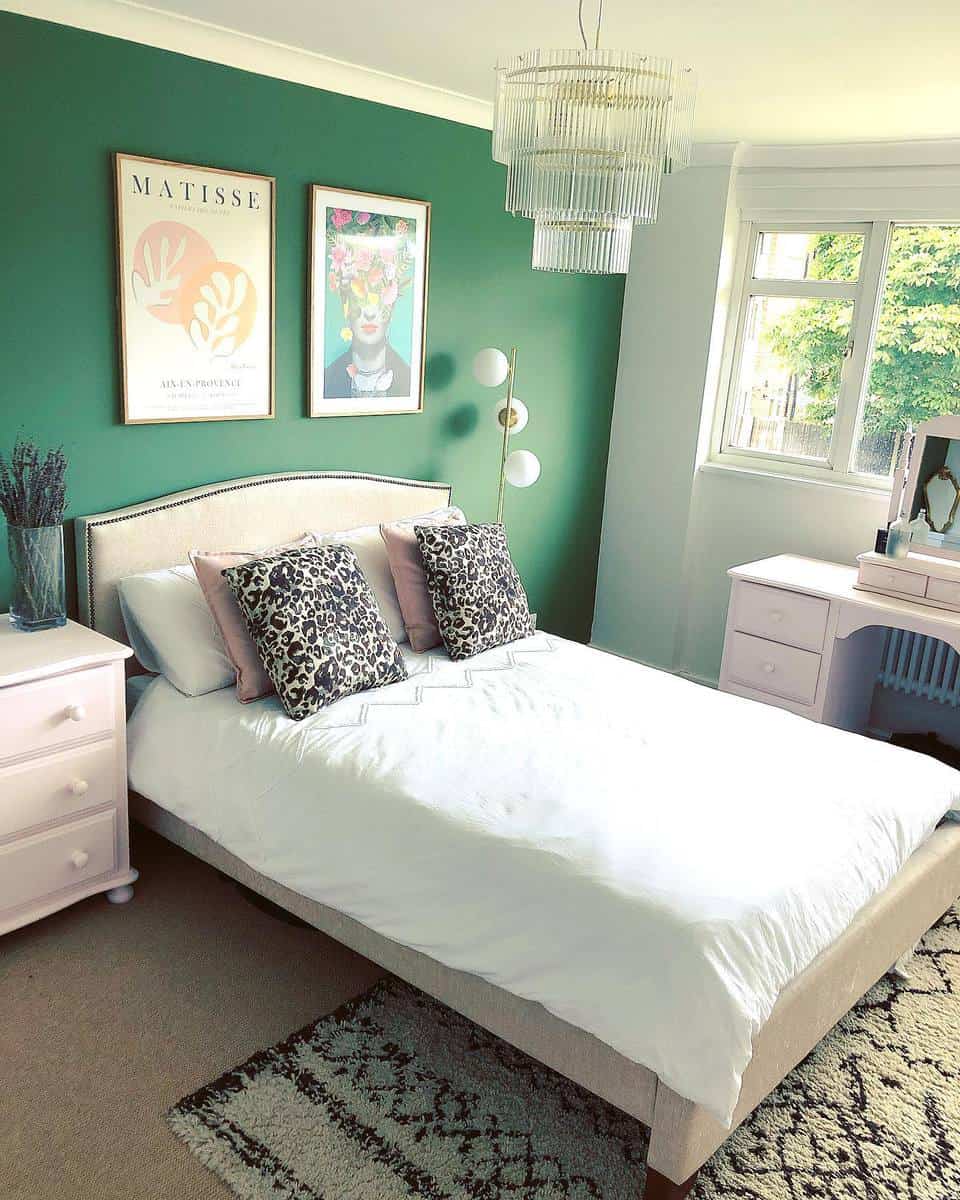 girl-green-bedroom-ideas-naomi-sian_-home_ Stylish bedroom with a green accent wall, artwork, a white bed, and a chandelier next to a window with a garden view