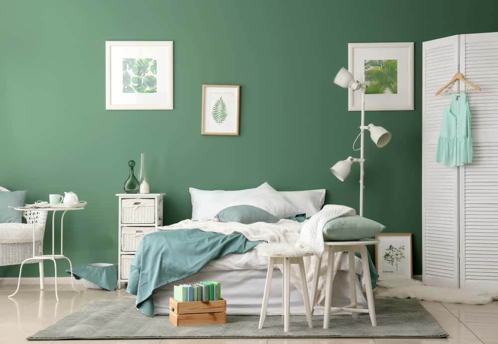 interior-green-bedroom-ideas-6 A green-themed bedroom with a bed, white lamp, and framed art on a green wall. A chair holds books and a cushion