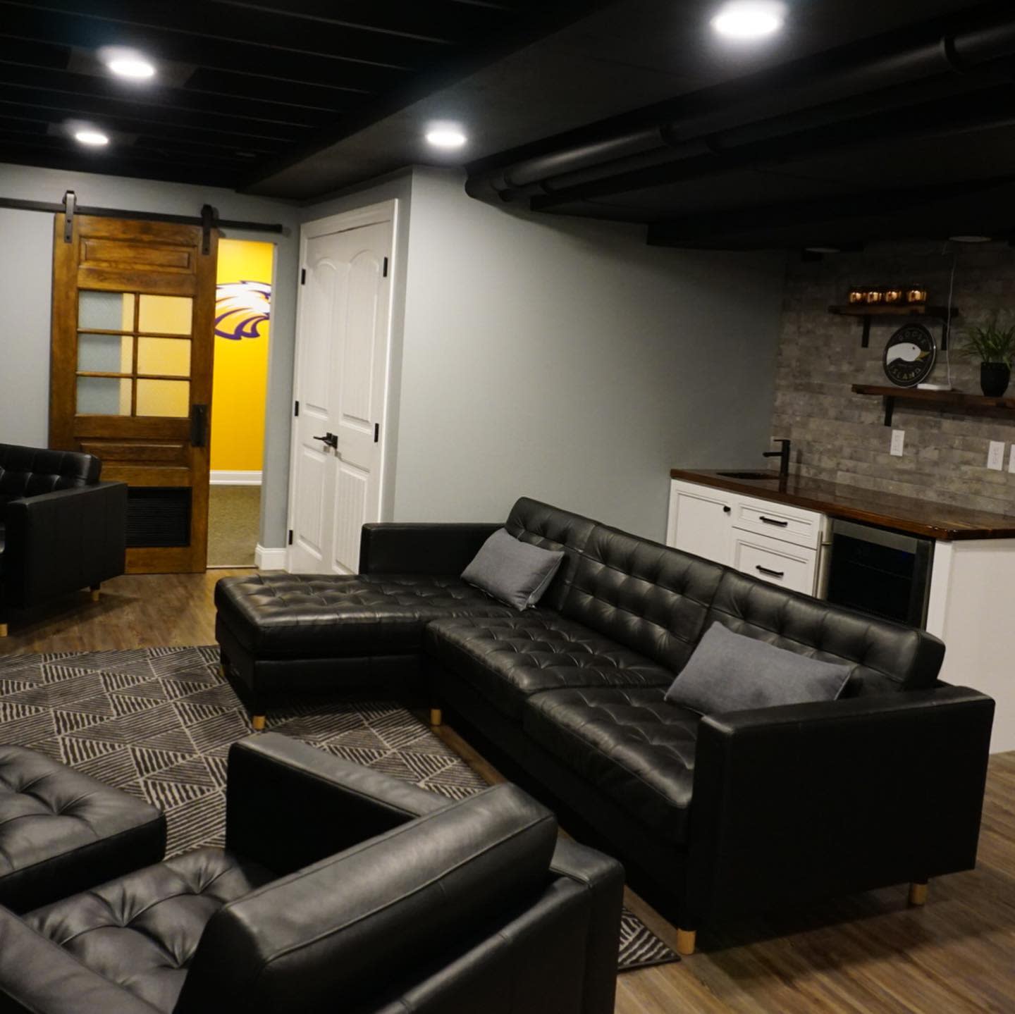 living20room20basement20apartment20ideas20-nhdconstructiondesign Basement living room with large black sectional, wood barn door, and small kitchenette.