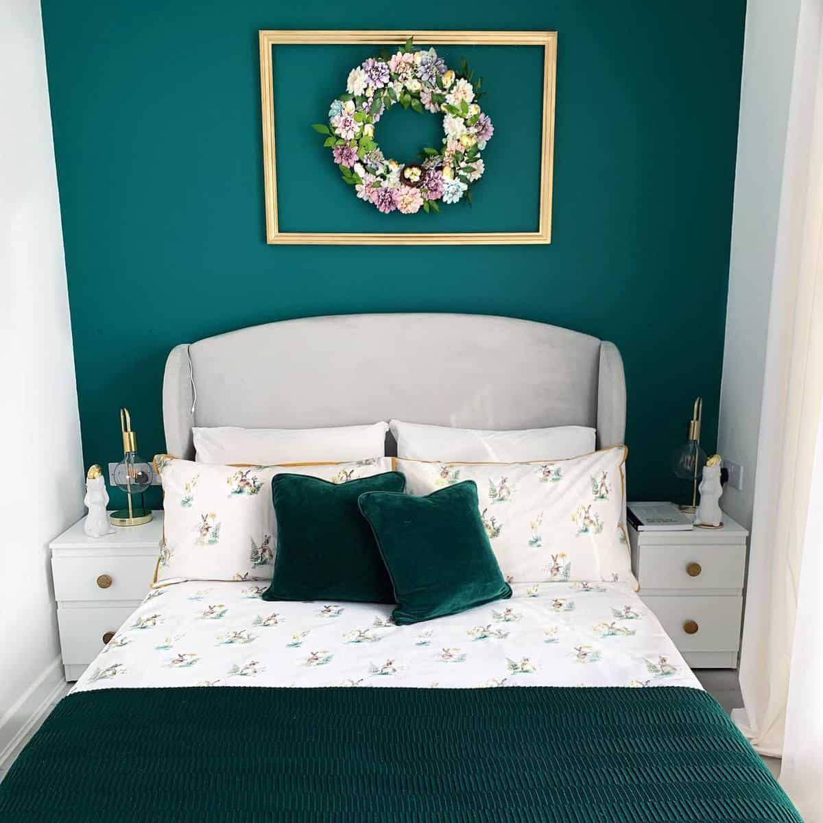 master-green-bedroom-ideas-cztery_katy_sylwi_ Cozy bedroom with teal accent wall, floral wreath, and a neatly made bed with patterned and solid green pillows