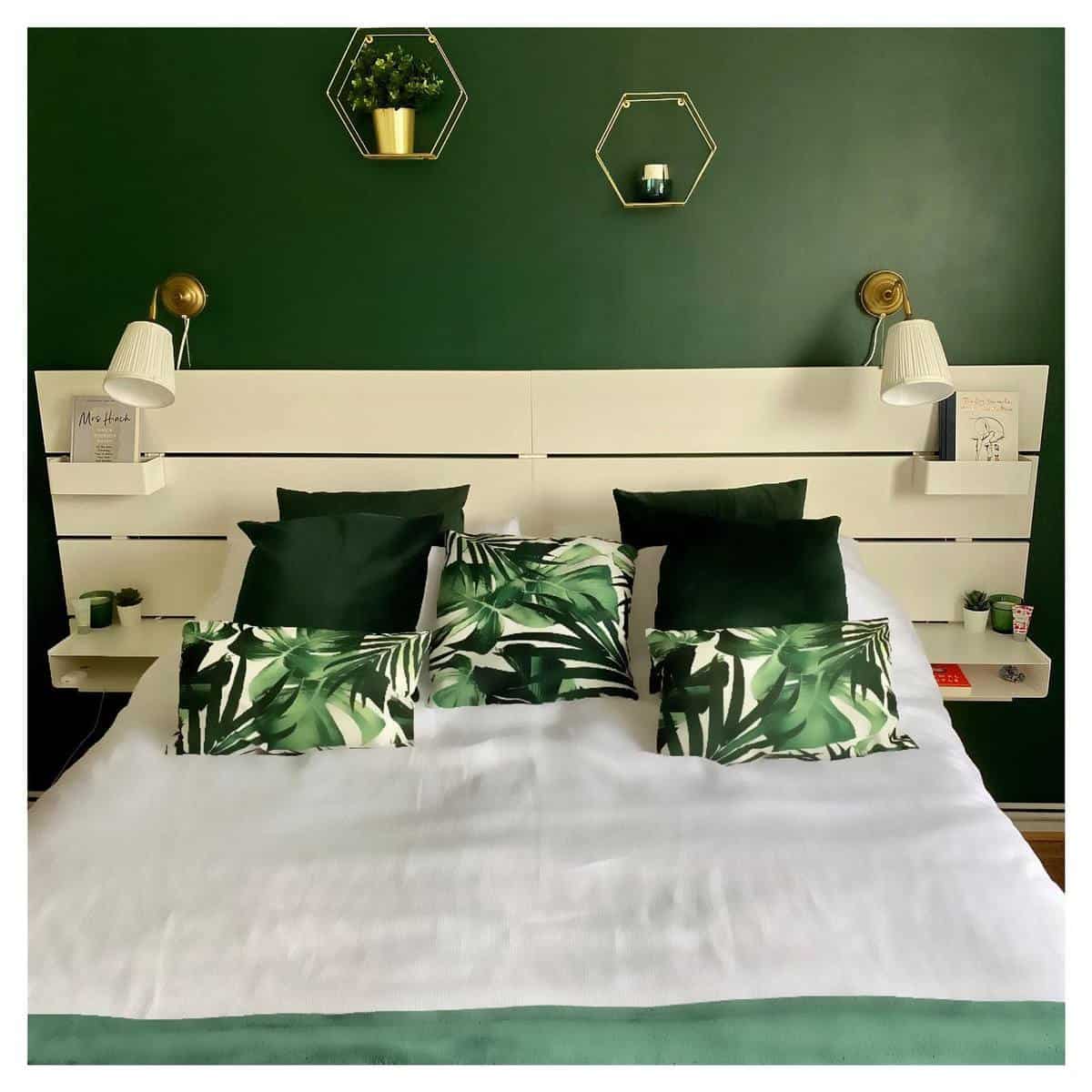 modern-green-bedroom-ideas-2-meandmyhome_alg A stylish bedroom with a white bed, green pillows, leaf-patterned cushions, and a dark green accent wall with decor