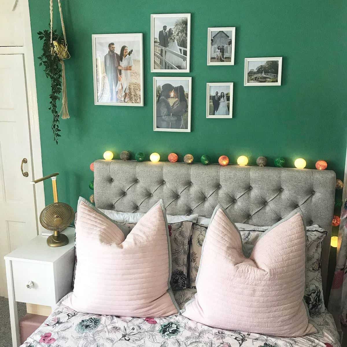 modern-green-bedroom-ideas-theahmedshome Bedroom with a gray tufted headboard, pink pillows, green wall with photos, and string lights above the bed