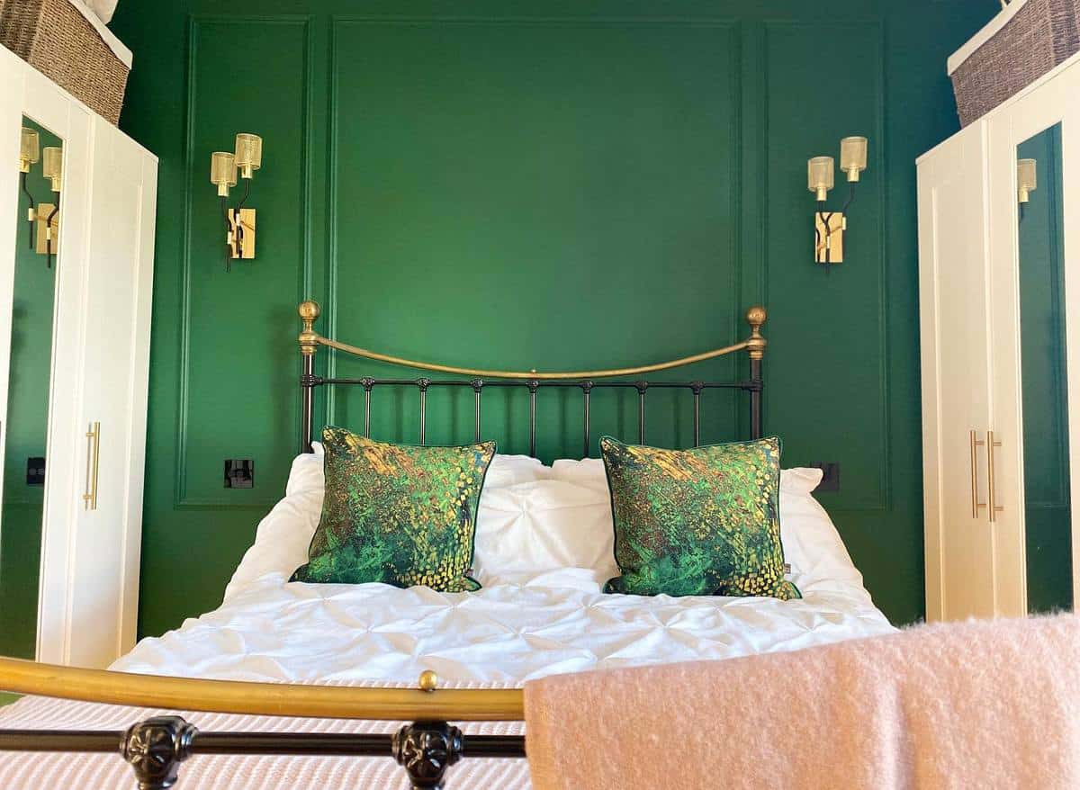 rustic-green-bedroom-ideas-canwepaintyet Cozy bedroom with a green accent wall, brass bed, and decorative pillows, flanked by white wardrobes and wall lamps