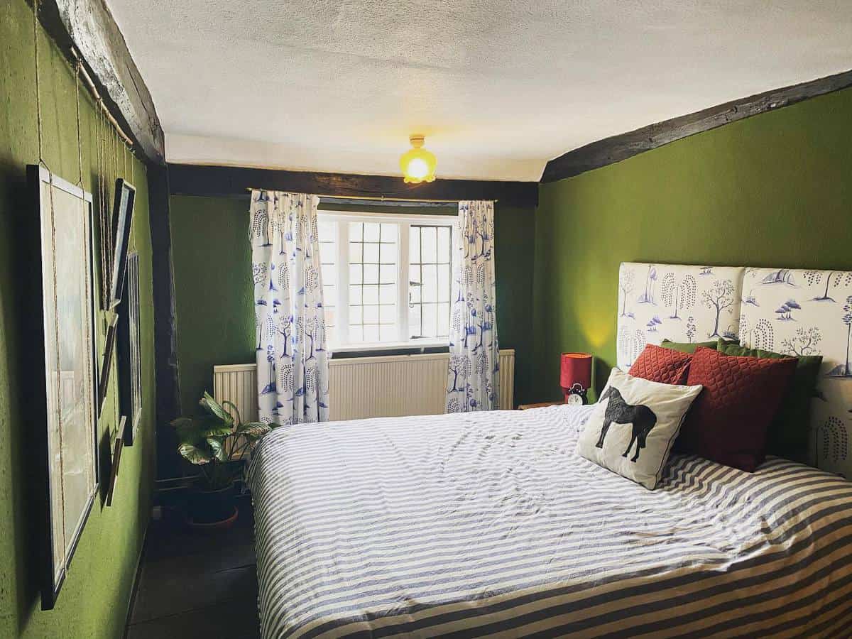 rustic-green-bedroom-ideas-creativelivingwithbarb Cozy bedroom with green walls, striped bedspread, horse-themed pillows, and floral curtains by a window