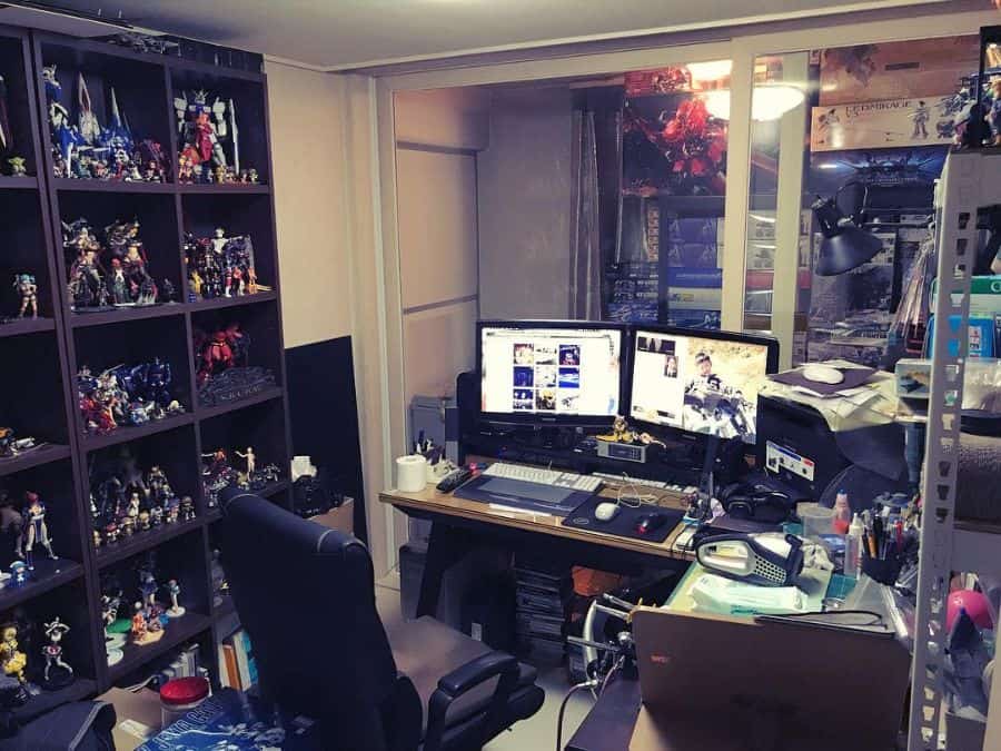 A cluttered home office with dual monitors, a gaming chair, shelves of figurines, and various electronics
