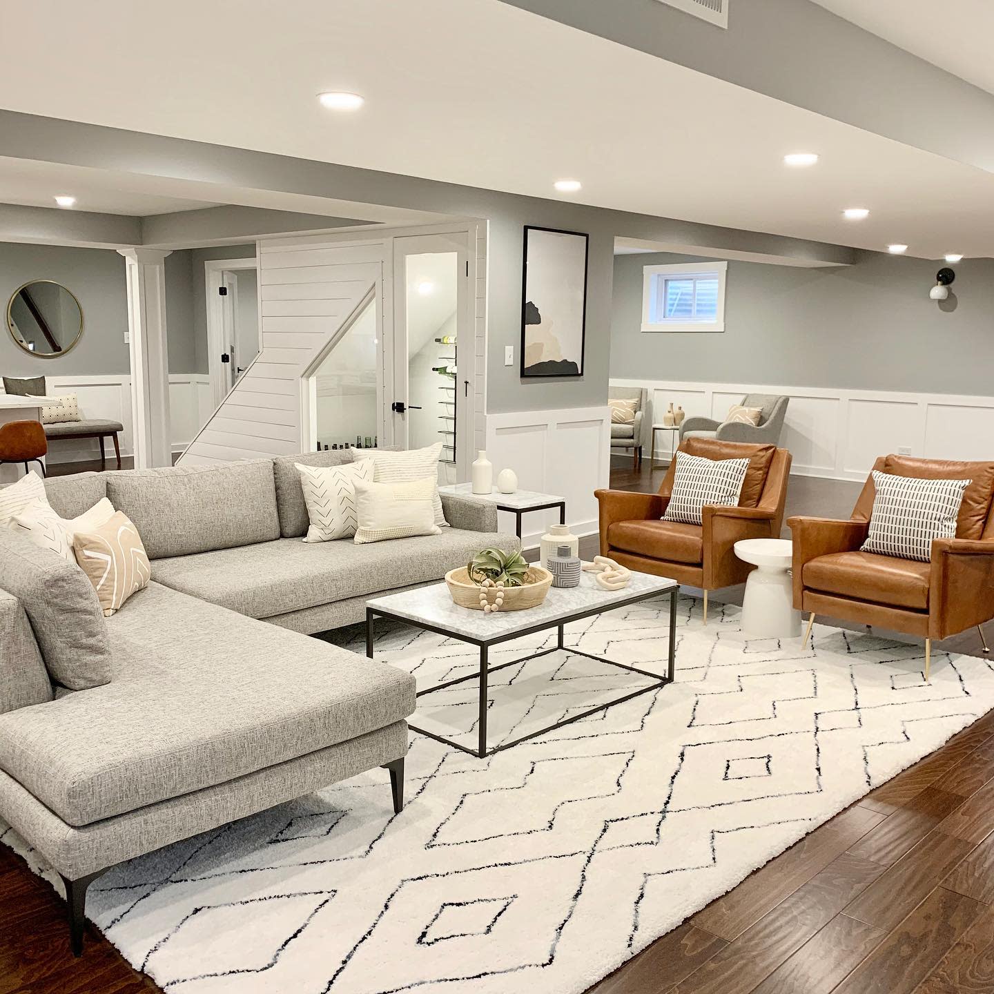 suite20basement20apartment20ideas20-whitebrassandmarble Modern basement living space with gray sectional, leather chairs, and patterned rug.